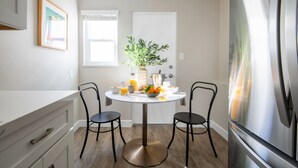 A modern kitchen with a white countertop, two black chairs, a round table with a potted plant, and a stainless steel refrigerator.