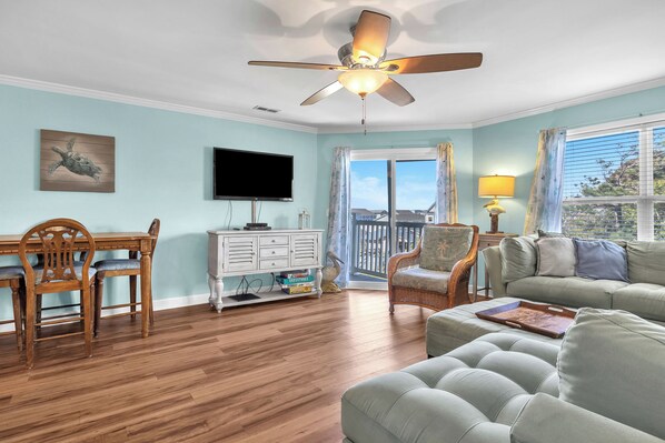 A living room with a wooden dining table, two chairs, a sofa, a ceiling fan, a television, and a window with curtains.