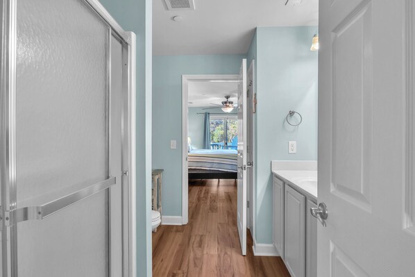 A hallway with a wooden floor leading to a bedroom with a bed and a window. - A Turtle's Pace: Ocean View, End Unit Condo in Walking Distance of Beach Access (Oak Island)