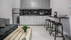 A compact kitchen with grey cabinets, a microwave, and a sink. There is a wooden bench with a plant, and a small table with stools. - Terra di Meteora Suites #3 (Kalampaka)