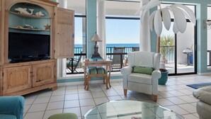 A spacious living room with a television, wooden cabinet, and a glass coffee table. - Gulf Front, Gorgeous Views, Top Floor, Pool, Gated Complex, 30A (Santa Rosa Beach)