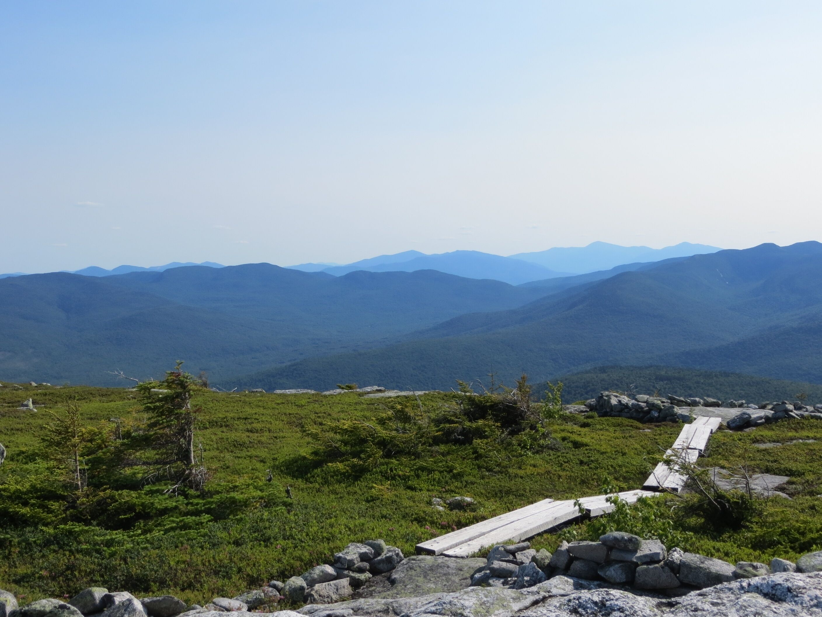 A mountain landscape with a wooden pathway leading through a grassy area with scattered rocks.