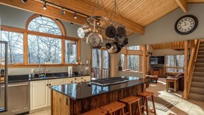 A spacious kitchen with a central island, wooden bar stools, large windows, and a hanging pot rack. - Beautiful Mountain Home - Next to Sunday River (Newry)