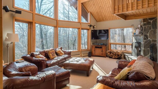 A cozy living room with leather sofas, a stone fireplace, and large windows offering a view of the forest. - Beautiful Mountain Home - Next to Sunday River (Newry)