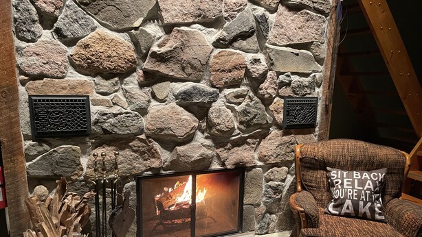 Une cheminée en pierre avec un feu de bois, un porte-bûches et un fauteuil confortable avec un coussin.