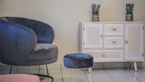 Une pièce comprenant un fauteuil gris, un petit pouf rond, une armoire blanche ornée de deux plantes en pot et un coussin rose.