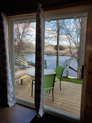 Een kamer met een grote schuifglazen deur die uitkomt op een terras met een tafel en stoelen, met uitzicht op een waterpartij en bomen.
