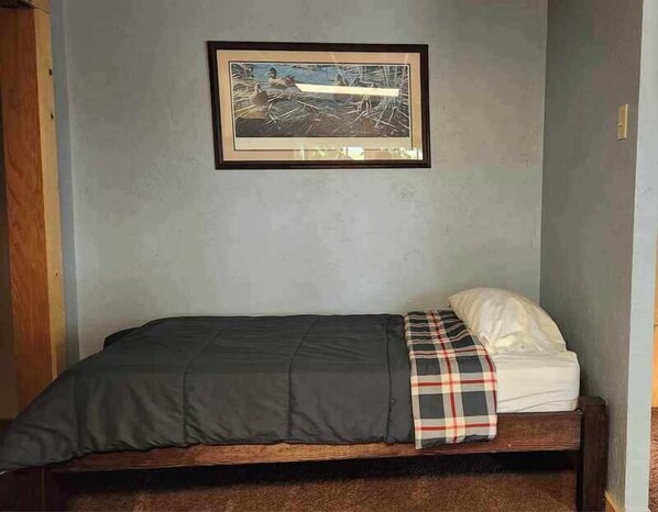 A single bed with a dark gray bedspread and a plaid blanket, a framed picture on the wall, and a wooden headboard.