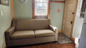 Una habitación pequeña y acogedora con un sofá color beige, una puerta de madera y una ventana con persianas.