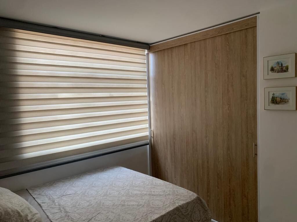 A bedroom with a bed, a window with blinds, a wooden wardrobe, and two framed pictures on the wall.