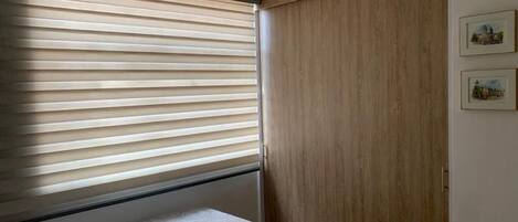 A bedroom with a bed, a window with blinds, a wooden wardrobe, and two framed pictures on the wall.
