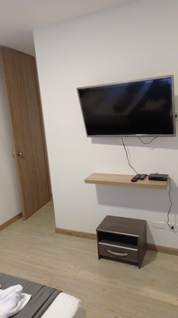 A room with a flat-screen TV mounted on the wall, a wooden shelf below it, and a small drawer unit.