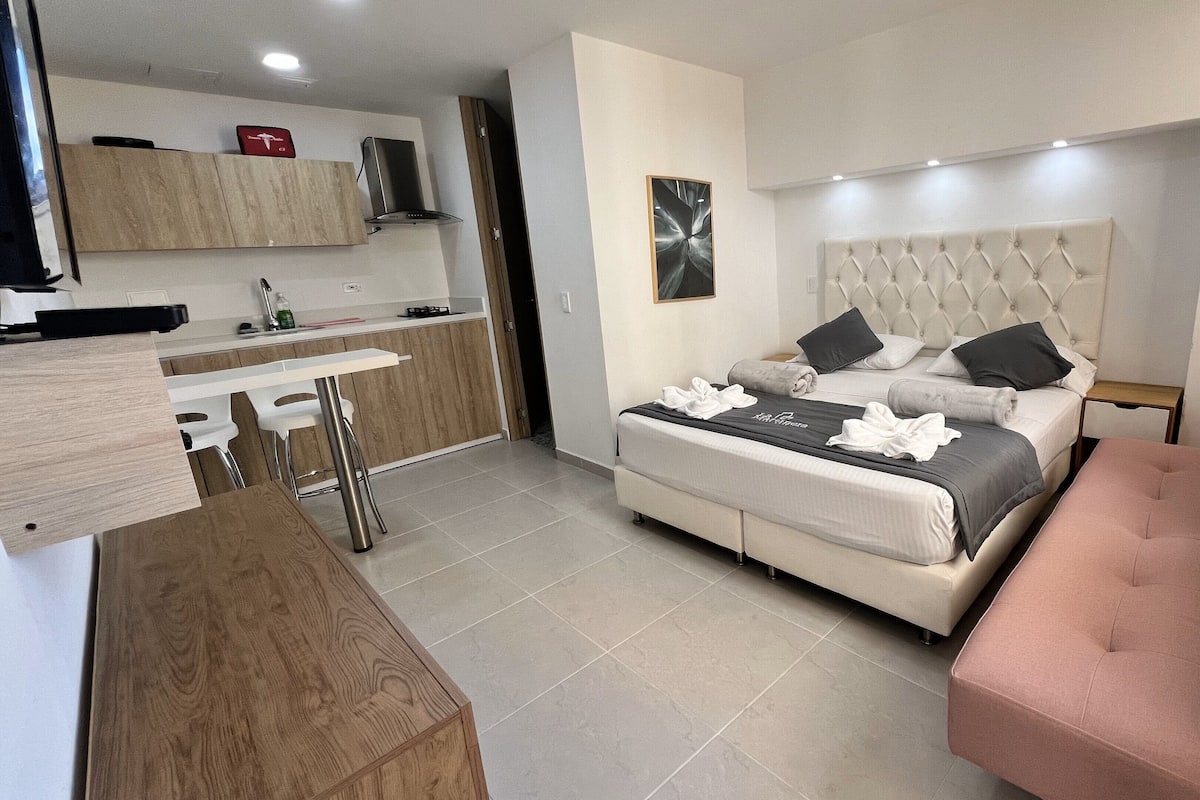Een moderne hotelkamer met een bed, een roze fauteuil, een kitchenette en een eettafel.