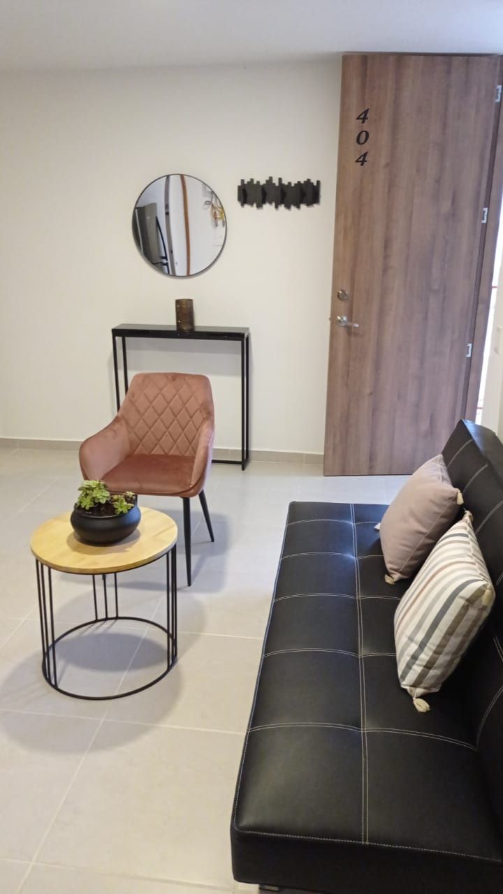 Een moderne woonkamer met een zwarte bank, een bruine fauteuil, een ronde salontafel met een plant, een spiegel aan de muur en een houten deur met het nummer 404.