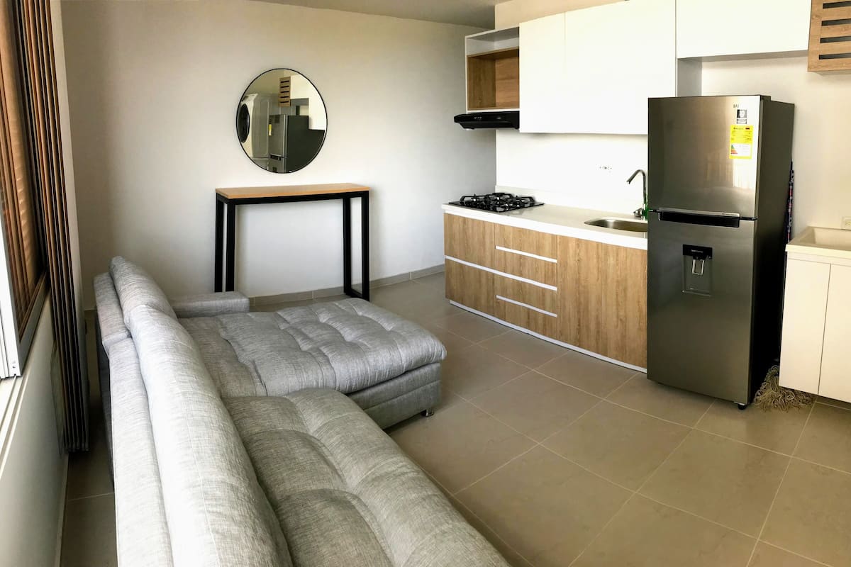 Een moderne keuken met een inbouwoven, een koelkast en een spoelbak. Er staat een grijze modulaire bank en een kleine tafel met een spiegel erboven.
