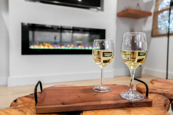 Two wine glasses on a wooden tray in a room with a fireplace and a television mounted on the wall.
