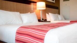 A hotel room with two beds, white bedding, a striped throw blanket, a nightstand, a lamp, and a framed picture on the wall. - Legends Casino Hotel (Toppenish)