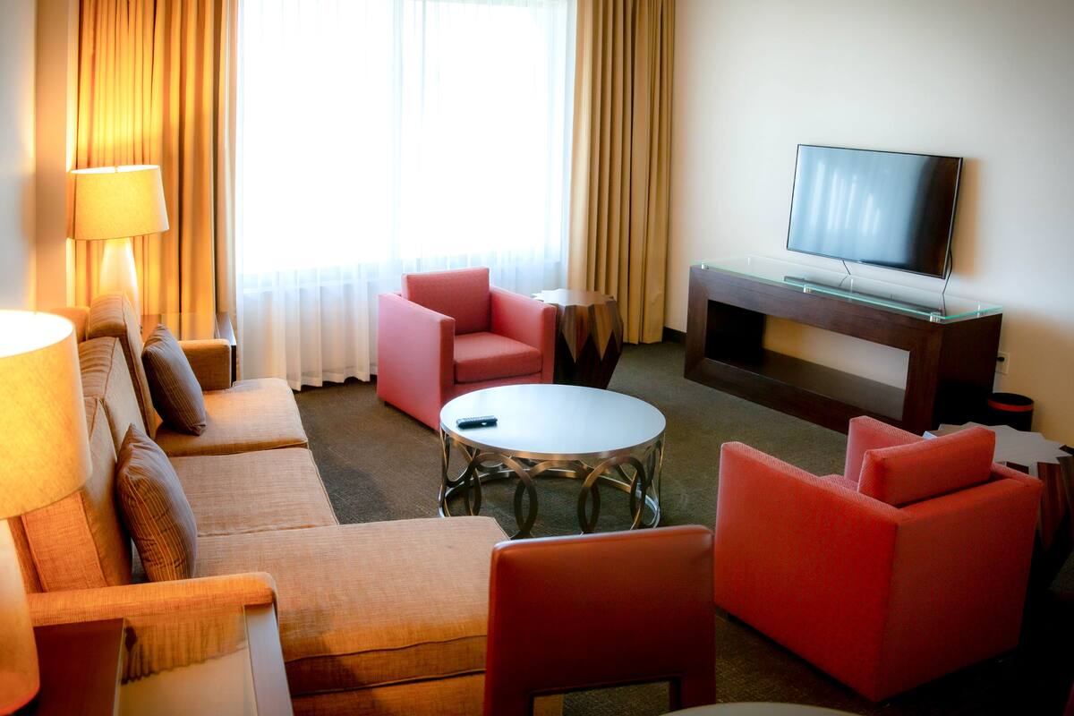 A hotel room with a sofa, two armchairs, a coffee table, a TV on a stand, and a lamp.