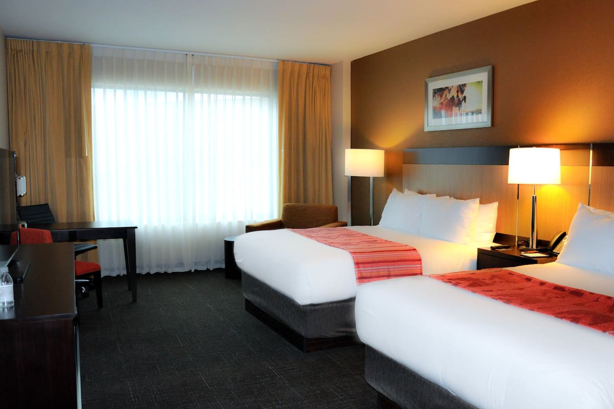A hotel room with two beds, a desk, a chair, and a nightstand with a lamp.