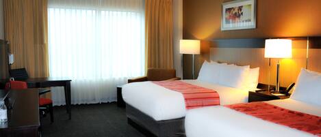 A hotel room with two beds, a desk, a chair, and a nightstand with a lamp.