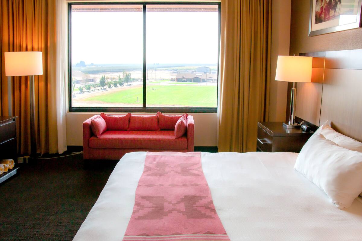 A hotel room with a large bed, a red sofa, a view of a field through a large window, and two bedside lamps.