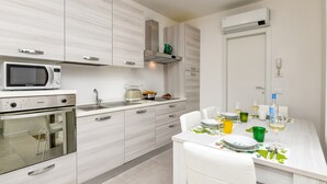 Eine moderne Küche mit weißen Schränken, einer eingebauten Mikrowelle und einem Backofen. Es gibt einen Essbereich mit einem Tisch, der für zwei Personen gedeckt ist, mit einem grünen Weinglas und einem weißen Weinkelch.