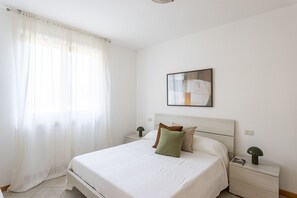 Quarto com cama, mesa de cabeceira, abajur, pintura e cortinas leves.