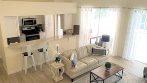 A modern living room with a sofa, coffee table, and chairs. A kitchen with white cabinets and appliances is visible in the background.