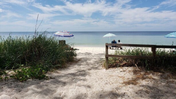 A sandy beach path leading to a wooden bench with two people sitting. Beach umbrellas are visible in the background.