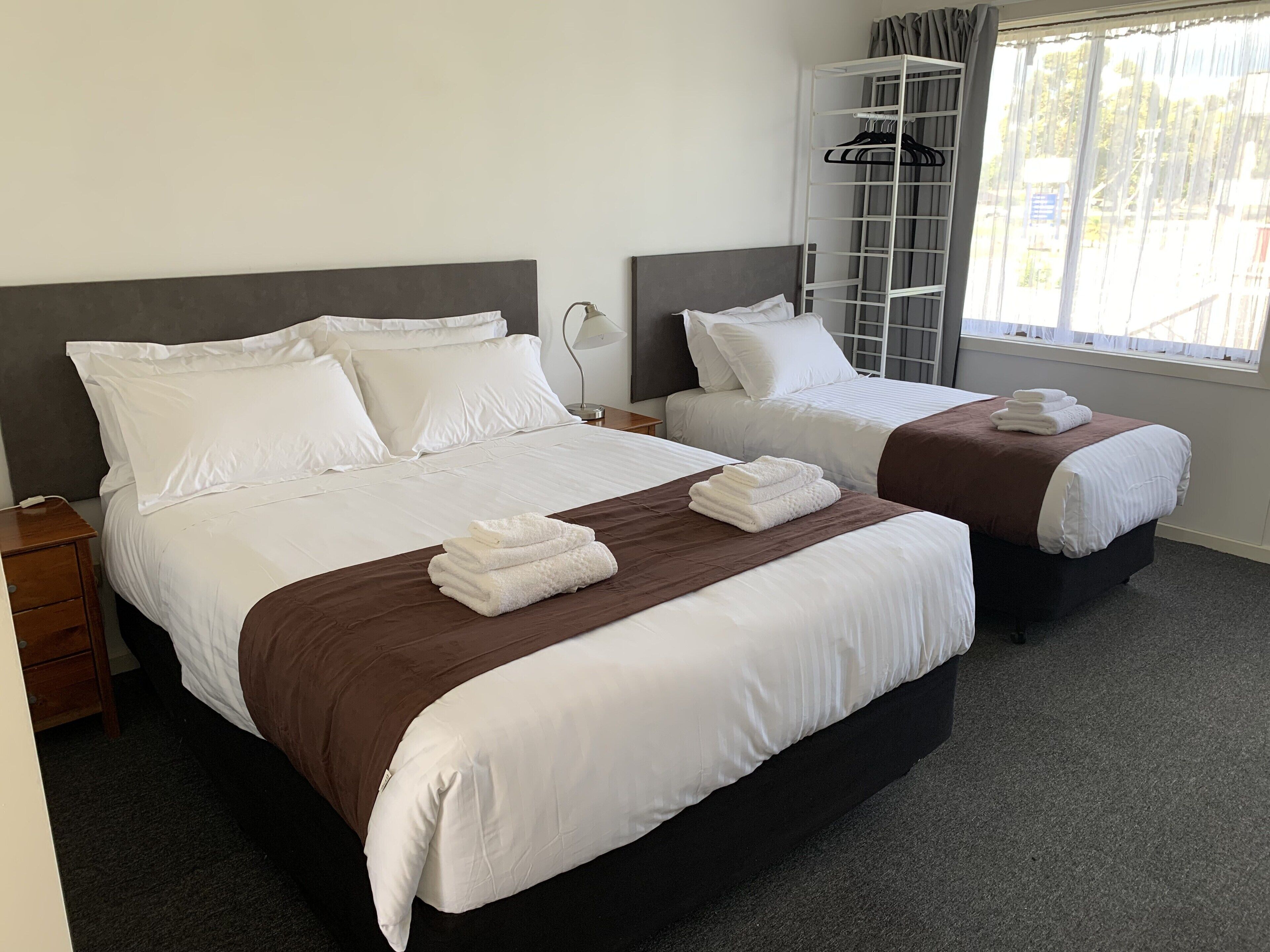 A hotel room with two beds, each with white linens and brown bedspreads. There is a white nightstand with a lamp and a small table beside the beds. A white wardrobe with a mirror is visible in the background.