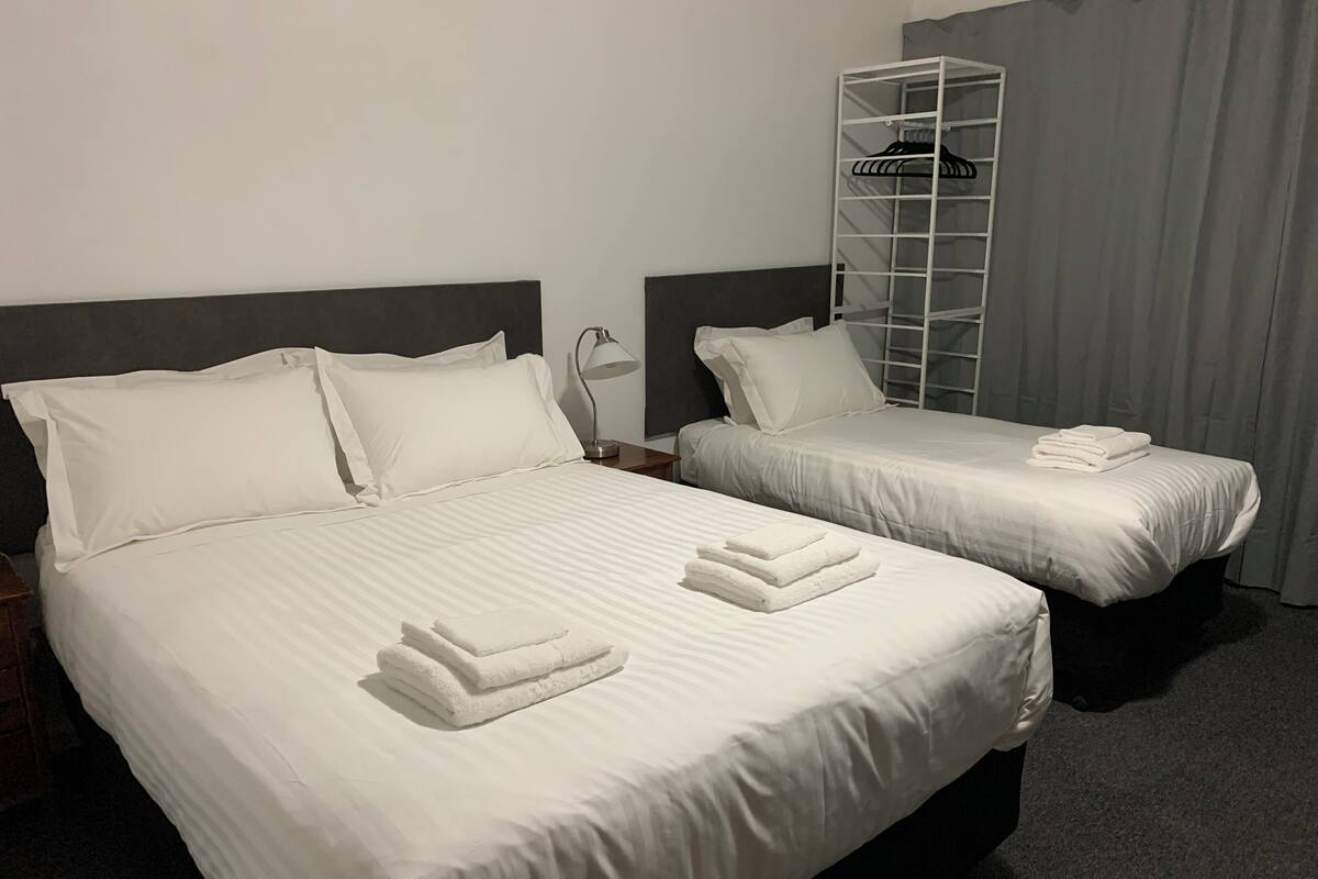 Two single beds with white linens and towels, a metal wardrobe, and a grey curtain.