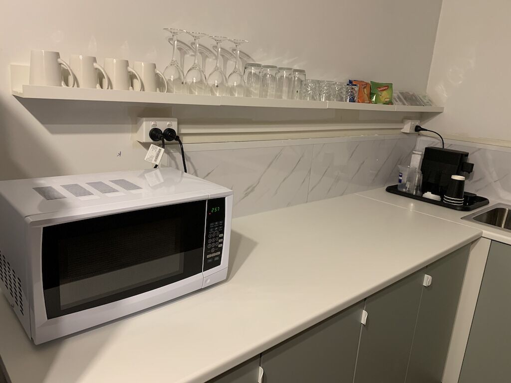 Una cocina con microondas blanco, cafetera y estante con vasos y tazas.