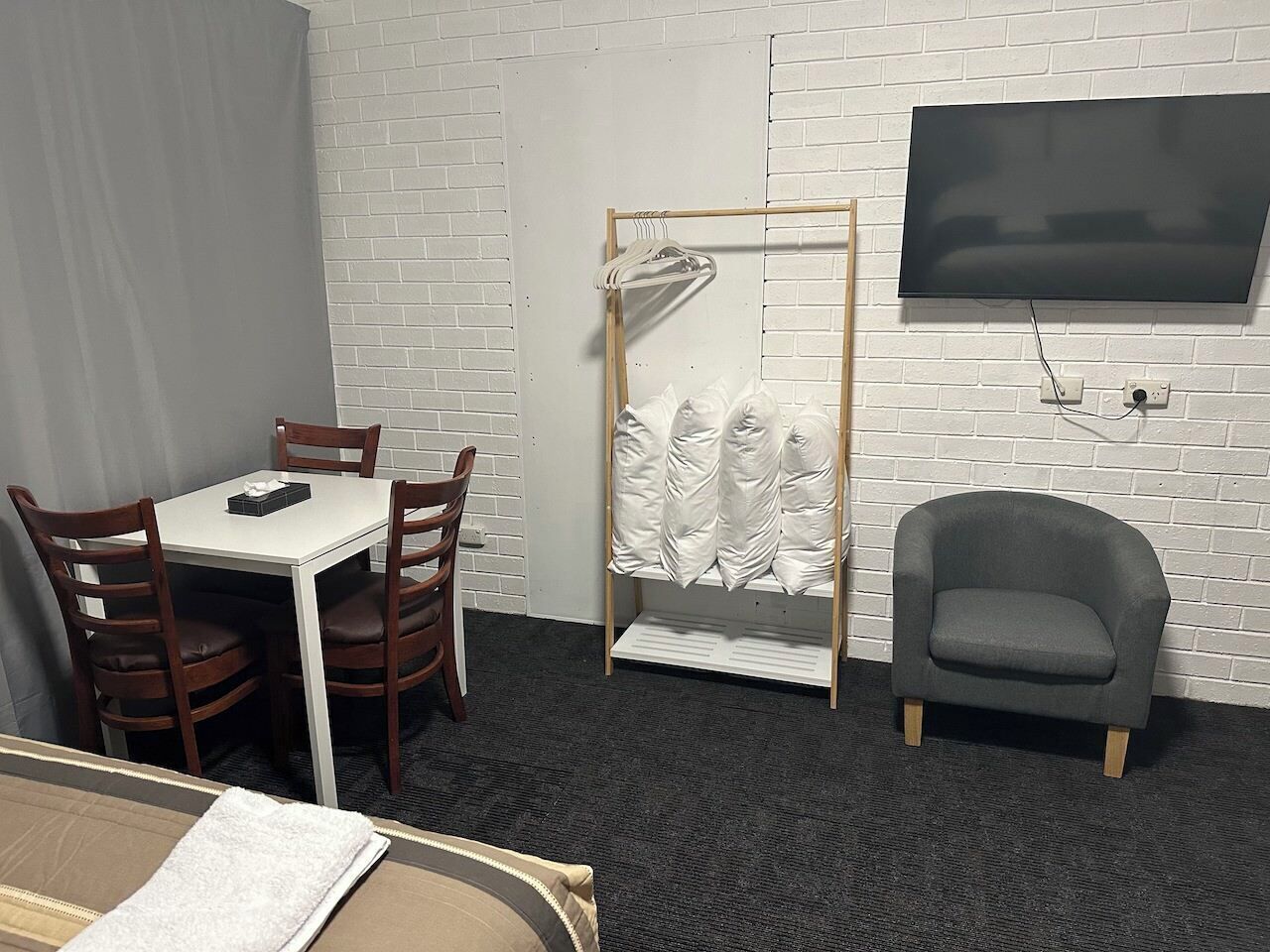 Une chambre d’hôtel avec un mur en briques blanches, un téléviseur à écran plat fixé au-dessus d’une chaise en bois, une petite table avec une chaise et un fauteuil gris.