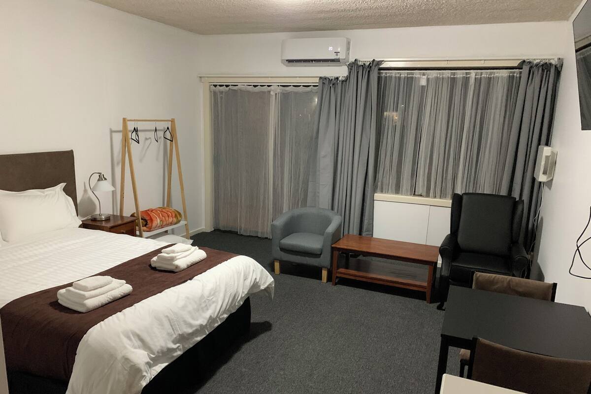 A hotel room with a bed, a desk, a chair, a small table, a lamp, a mirror, and curtains.