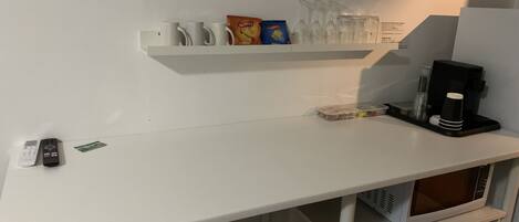 A white counter with a coffee machine, a microwave, and a shelf with cups, a box of Sprinters, and a bag of snacks.