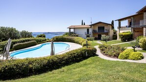 A pool surrounded by well-maintained hedges, a clear view of the sea, and a two-story building with balconies.