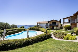 A pool surrounded by well-maintained hedges, a clear view of the sea, and a two-story building with balconies.