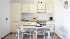 A compact kitchen with white cabinets, a dining table with four chairs, a coffee maker, and a small potted plant.