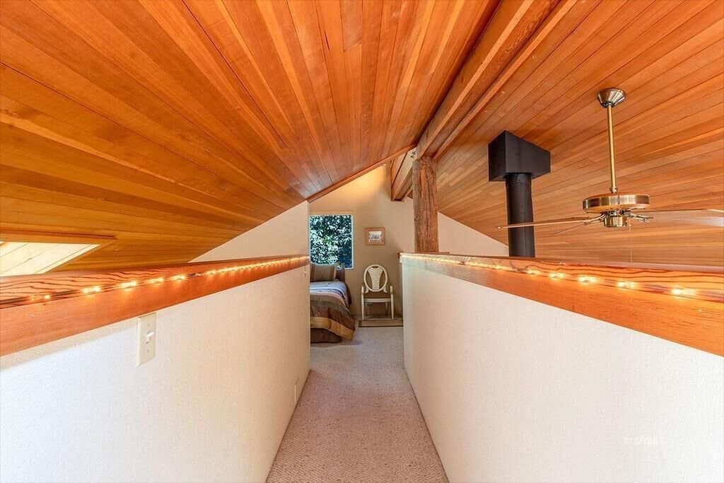 A narrow attic room with wooden beams, a ceiling fan, and a bed with a view of trees outside.