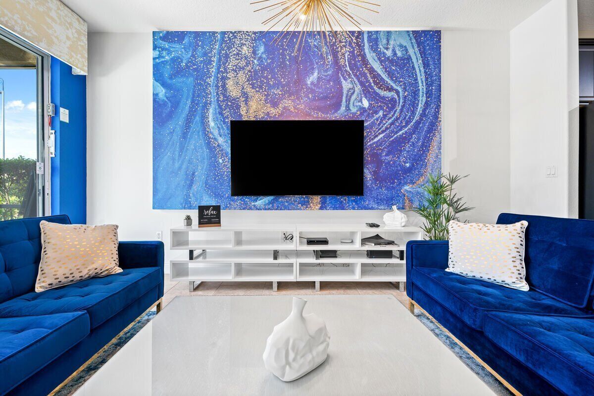 A modern living room with a large flat-screen TV mounted on a white wall, flanked by two blue sofas and a white coffee table with a decorative vase.