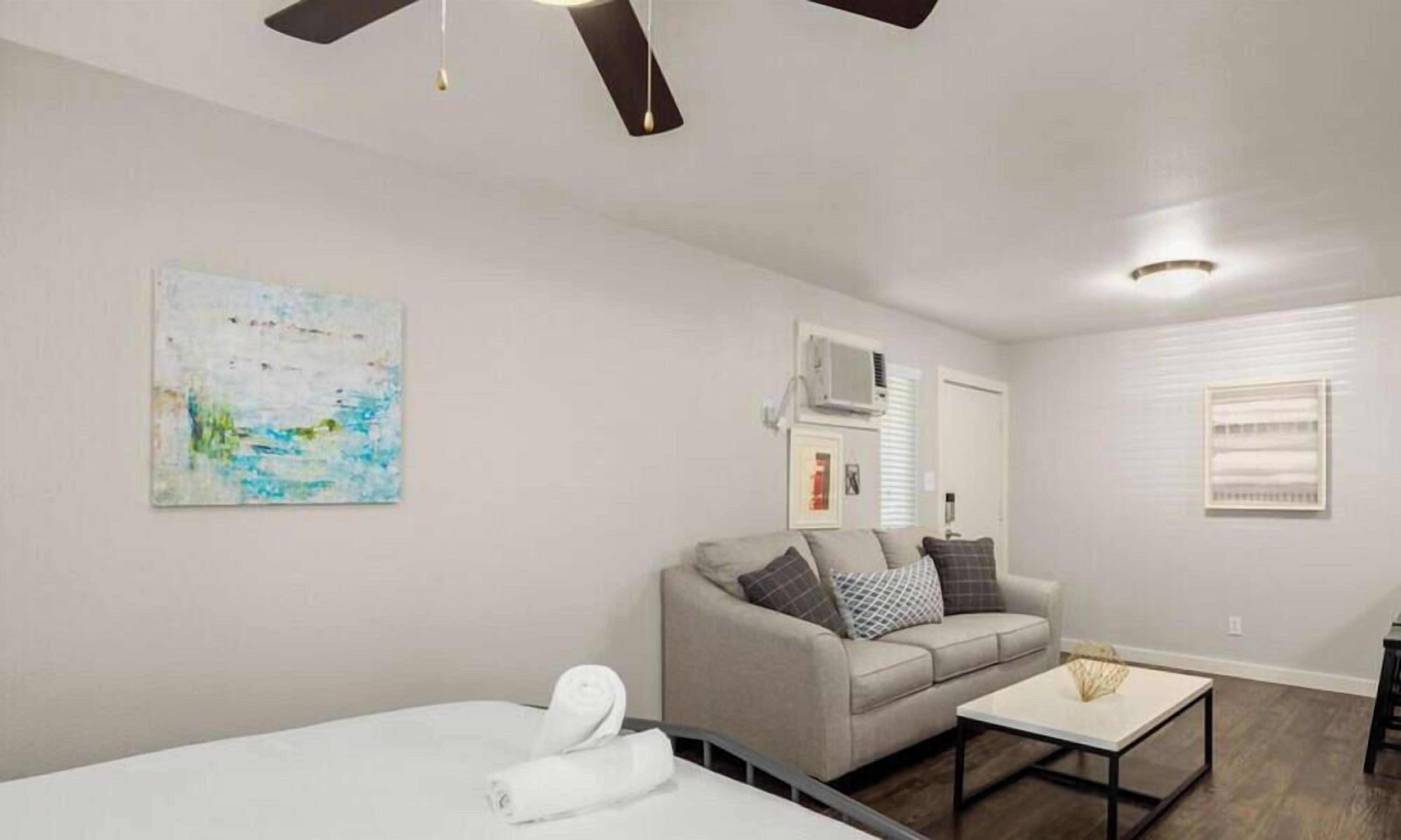 Un soggiorno moderno con un letto grande, un divano e un tavolino. C'è una ventola a soffitto, un quadro al muro e una finestra con persiane.