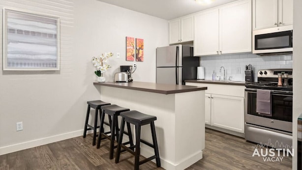 A modern kitchen with white cabinets, a stainless steel refrigerator, a microwave, and a gas stove. There are three bar stools and a small island with a sink. - HC #102 xxxxxxxxxxxx (Austin)
