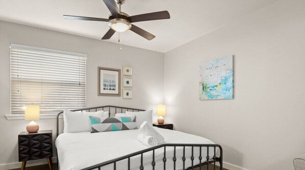 A bedroom with a bed, a ceiling fan, a window with blinds, two bedside tables with lamps, and a wall with framed pictures and a painting.