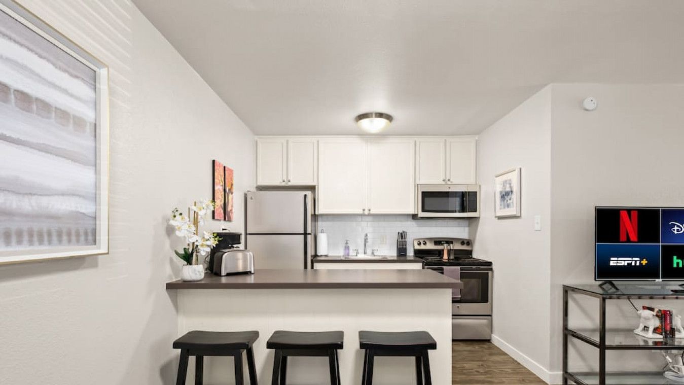 A modern kitchen with white cabinets, a stainless steel refrigerator, a microwave, and a stove. There are three black stools at the kitchen island.