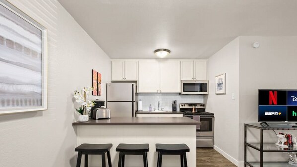 Una cocina moderna con armarios blancos, un refrigerador de acero inoxidable, un microondas y una estufa. Hay tres taburetes negros en la isla de la cocina.