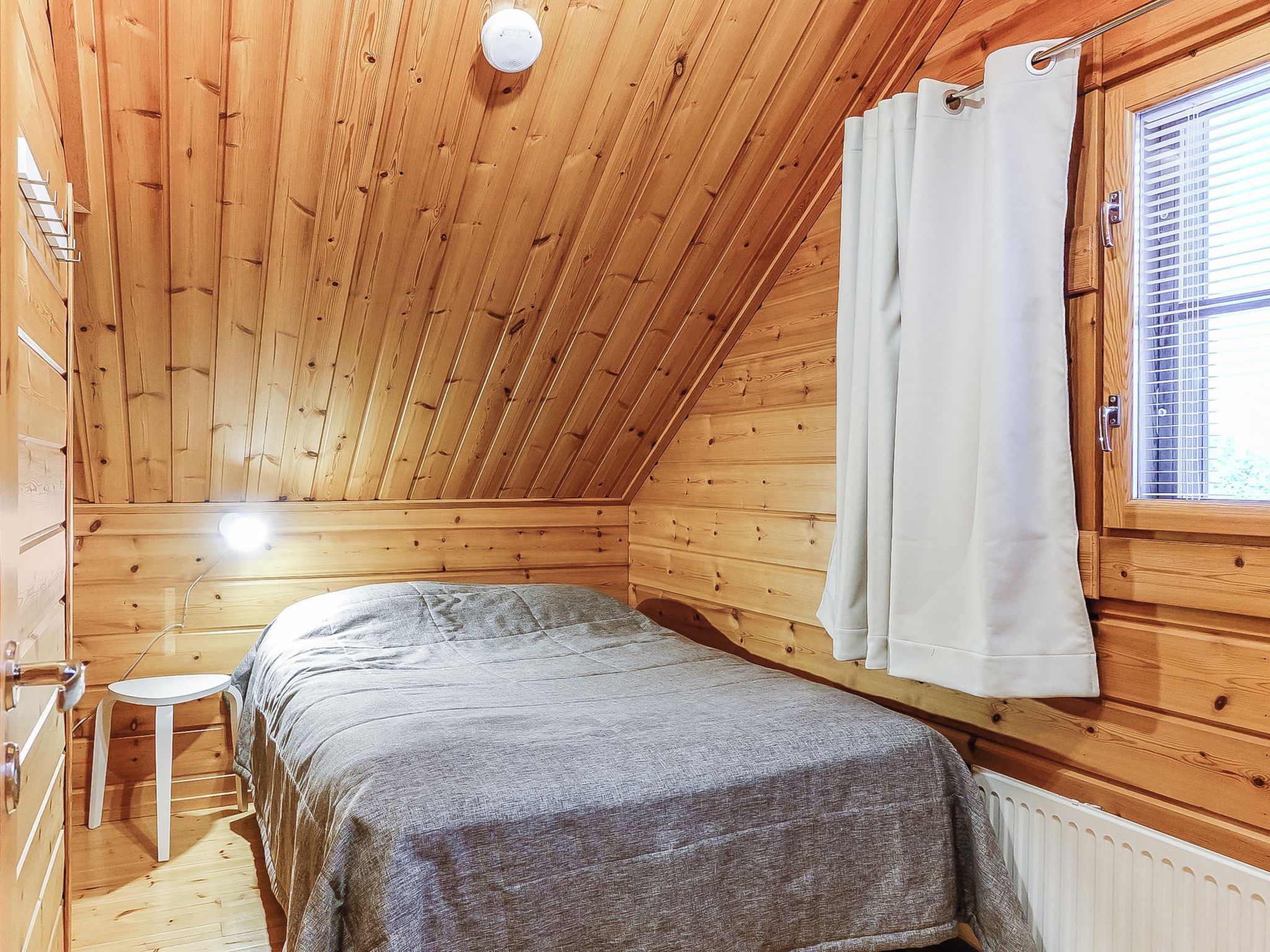 Et lille, hyggeligt rum med trævægge og et skråt loft. Der er en seng med et gråt tæppe, et lille hvidt bord og et vindue med hvide gardiner.
