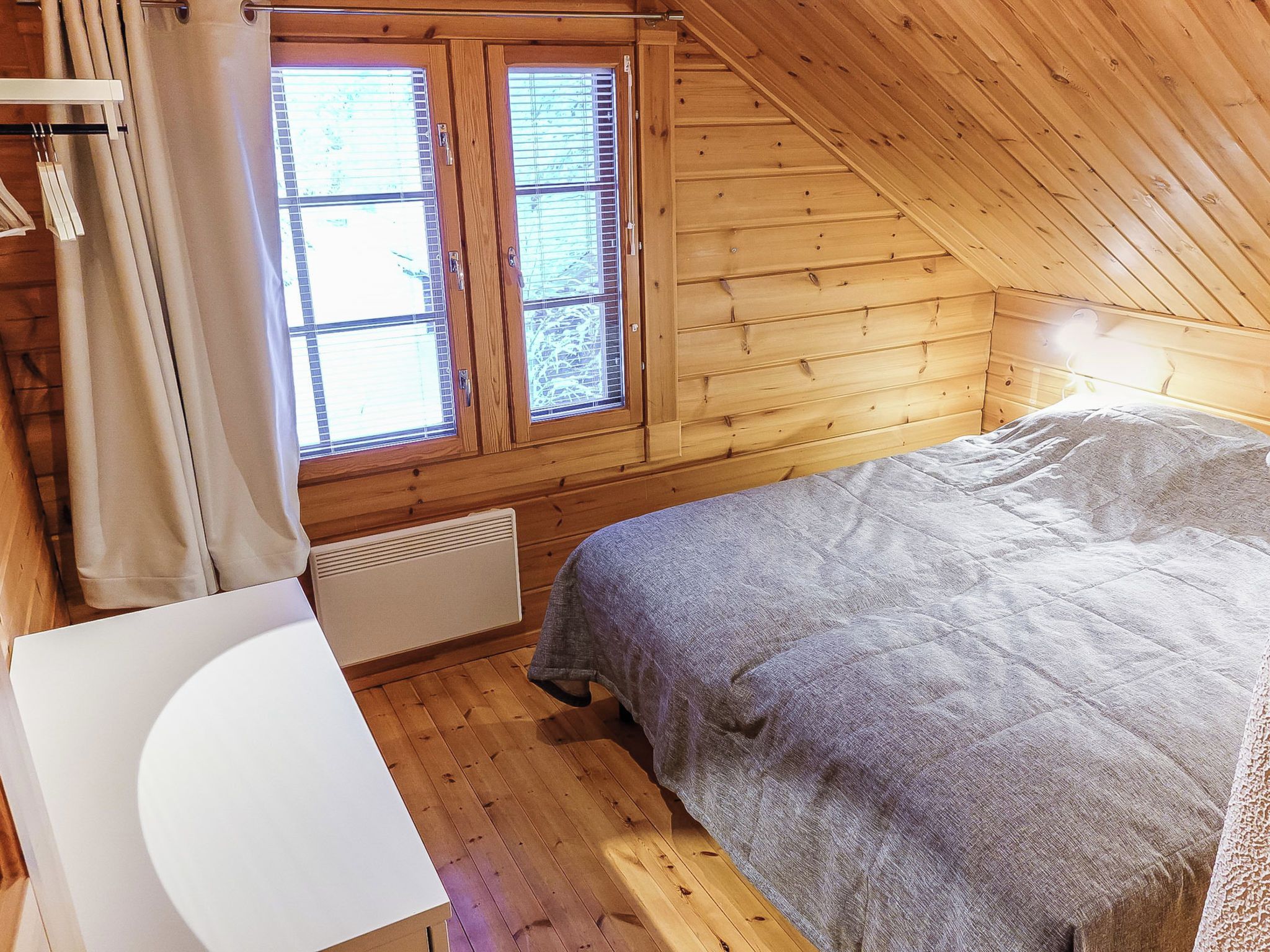 A bedroom with a sloped ceiling, wooden walls, a bed with a grey blanket, a white chair, and a window with blinds.