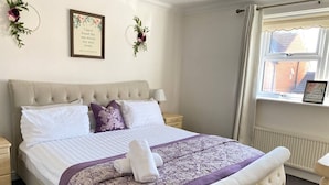 A neatly made bed with a patterned bedspread, two wall decorations, and a window with a view of a building. - 4-bed House With Private Garden in Barnsley! (Barnsley)