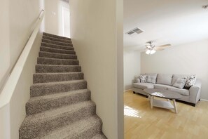 Uma escada com degraus revestidos de carpete conduz a um patamar, onde se encontram um ventilador de teto, luminária, um sofá com almofadas e uma mesa de centro.