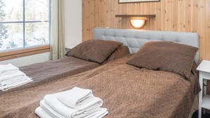 A double bed with brown bedding and pillows, a white nightstand, a wall-mounted lamp, and a window with blinds.